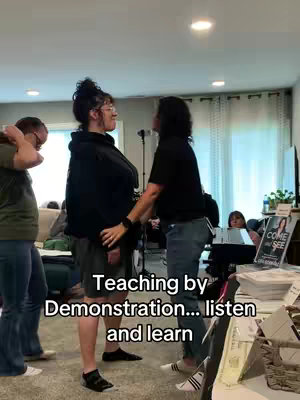 The image shows three women in what appears to be a living room setting. The central focus is a woman in a black hoodie and shorts, being guided by another woman who is wearing black. A third woman stands to the side with her back to the camera. The scene suggests teaching or demonstration, with the camera angle centered on the interaction between the women. The background is blurred, filled with indoor elements like a table, chairs, and window, suggesting a typical home environment. The color palette is neutral, featuring earth tones and muted shades.