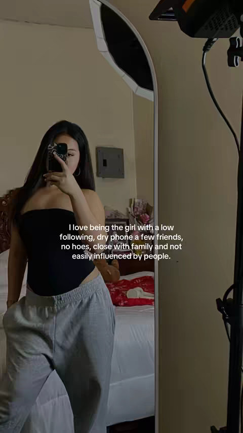 A young woman stands in front of a mirror, holding up her phone to take a selfie. She's wearing a black tube top and gray sweatpants. The room is softly lit, and some text is overlaid on the image.