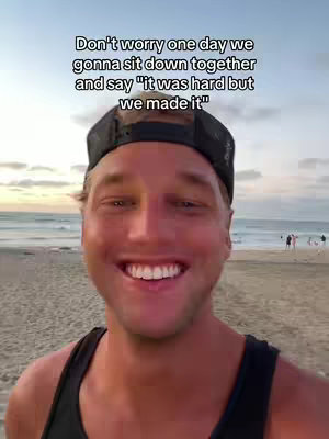 A selfie-style shot of a man smiling broadly at the viewer. He is wearing a black cap and a dark tank top. The background shows a beach with the ocean and a sunset sky, with text overlaid at the top.