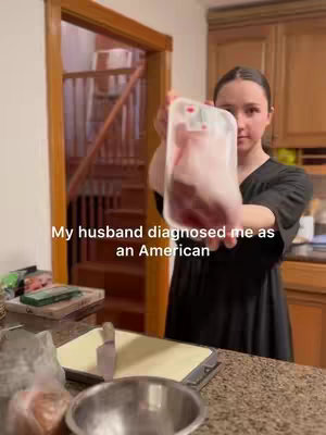 A woman in a black top is holding a package of meat in a kitchen. The background includes wooden cabinetry and a glimpse of a staircase. Text overlayed on the image reads 'My husband diagnosed me as an American'.