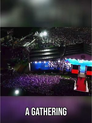 An aerial night shot shows a large outdoor gathering illuminated by bright stage lights and spotlights. A stage with a red carpet and a large screen is in the center, and a huge crowd of people are standing in front of it with their hands in the air. The sky is dark, and the image is framed by a purple banner with white text at the bottom that reads 'A GATHERING'.