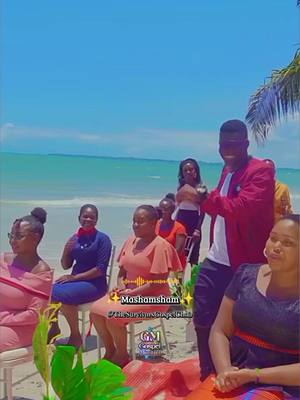 A group of people, including several women and a man, are gathered on a beach, possibly for an event. The man, dressed in a red blazer, appears to be addressing the group. The setting is a sunny beach with a clear blue sky and ocean in the background. Text overlay is present on image.
