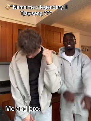 Two young men are in a kitchen setting. The individual on the left is dressed in a light grey zip-up hoodie and black shirt. The subject on the right is wearing a grey sweat suit. Above the left subject are dark brown cabinets, and behind them, simple off-white walls. A text overlay at the top frames the composition and the content.