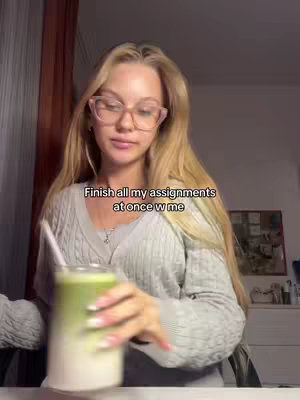 A young woman with long blonde hair and glasses is seated at a desk, holding a green drink in a clear glass. Text overlay reads, "Finish all my assignments at once w me."