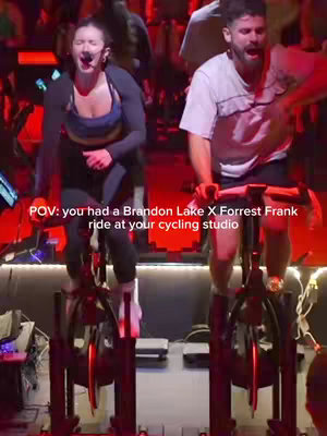 The cover image shows two people on stationary bicycles in a cycling studio, engaged in what appears to be a high-energy workout. The person on the left, a woman, is wearing a dark top with a sports bra and is singing into a microphone. The person on the right, a man, is also singing, and appears to be working really hard. In the image, the people and lights seems to give more energy to the video.
