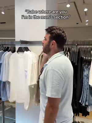 A man stands in a clothing store, facing away from the camera and looking towards a rack of clothing. Text is overlaid at the top of the frame.