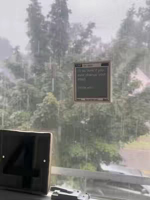 The image shows a window with rain falling outside. A small, retro-looking device displaying text is attached to the window. The background is blurred, with trees visible. In the bottom left corner, a dark object, possibly a part of the window frame or a device, is present.