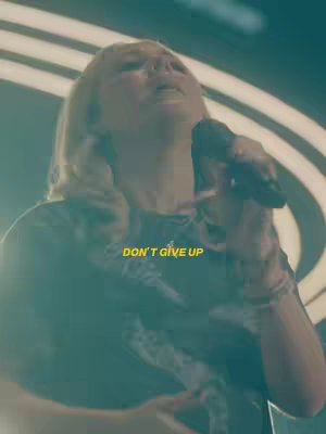 The image features a woman with blonde hair holding a microphone, singing. The text \"DON'T GIVE UP\"  is overlaid in a yellow font. The background seems to be a stage setting with lights.