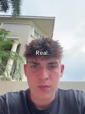 A young man with short, spiky hair is the primary focus, looking directly at the camera. The background is a blurred setting with a house and some greenery. Overlaid on the man's forehead is the text 'Real.'