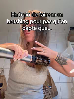 A woman is shown in a bathroom, holding a hairdryer and brush to style her hair. The text overlay suggests she is trying to not let on that something is happening.