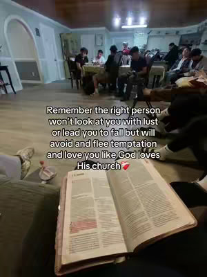 The image shows an open Bible on a person's lap. In the background, a social gathering appears to be taking place, with several individuals seated around a table. The room is lit with overhead lighting.