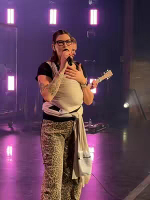 A woman wearing glasses, a black top, and patterned pants is holding a microphone and a small ukulele, standing on a stage with low lighting, likely in a concert performance. She is wearing a baby carrier and holding the microphone close to her mouth.