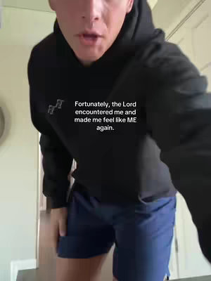 A man wearing a black hoodie and navy shorts stands in what appears to be a doorway, looking towards the camera. Text is overlaid on the image.