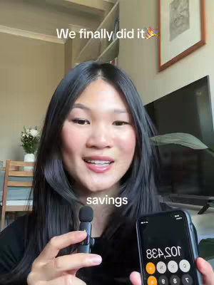 A woman with long dark hair stands in front of a light-colored wall, holding a microphone and a smartphone showing a calculator app. The text overlay says "We finally did it". The background includes a framed picture and a vase of white flowers.