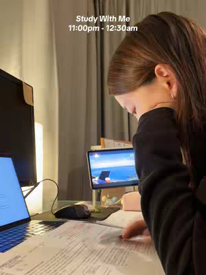 A young woman intensely studies at a desk. Her face is partially obscured as she leans over a textbook, highlighting the intensity of her studies. A laptop displaying a beach scene sits open on the desk, along with a notebook and a writing utensil. A lamp is visible, casting a warm light.