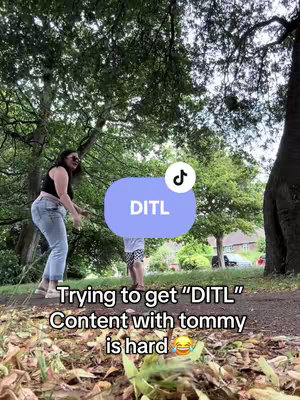 A woman in sunglasses, black top, and jeans stands in a park, interacting with someone out-of-frame. A light blue oval with the text "DITL" and a TikTok logo is in the middle, and text at the bottom of the frame reads "Trying to get "DITL" Content with Tommy is hard 😂."