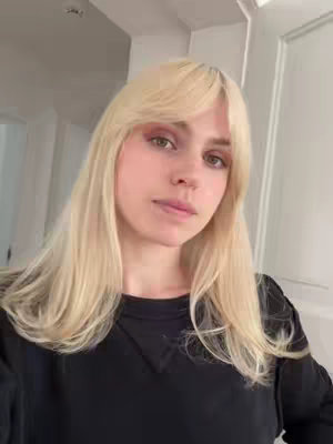 A selfie-style shot of a young woman with long, light-blonde hair that has a side part and bangs. She appears to be in a well-lit room, and her eyes are directed towards the camera. She is wearing a black crew-neck shirt. The background is blurred, but appears to include a white wall and doorframe.