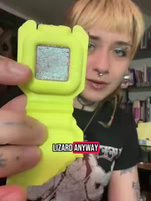 A person with light skin and short, light blonde hair angles the camera towards themselves, holding a bright yellow piece of packaging. The package sits close to the camera, featuring a rectangular section holding a product with a reflective surface that appears to be eyeshadow. The person is wearing a black t-shirt and has multiple facial piercings.