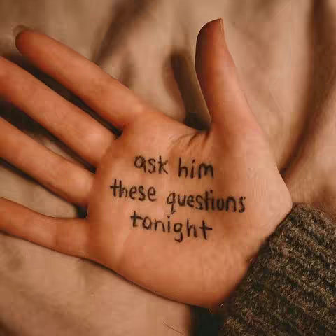 A close-up of a light-skinned hand with text written on the palm. The hand is slightly open, and the text says, "ask him these questions tonight." The background has a soft, blurred, warm-toned fabric.