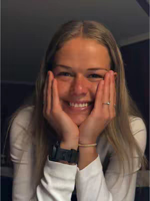 A close-up shot of a young woman with blonde hair, holding her hands up to her face with a smile. She's wearing a white long-sleeved shirt and a watch. The background is dark.