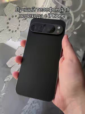 A hand holds a black smartphone with a unique camera module aligned to the left. The background reveals a patterned, slightly blurred curtain. The image composition is simple, focusing on the phone and the hand.