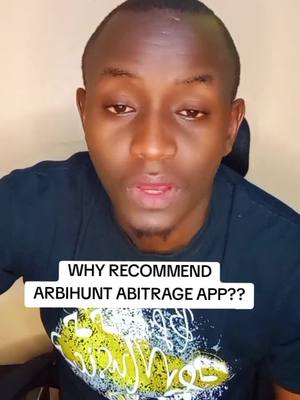 A man is centered in the frame, facing the camera and speaking. He has short dark hair and is wearing a dark blue t-shirt with a graphic design that is partially visible. The background is a light, neutral color. A white rectangular overlay at the bottom of the image contains the text, 'WHY RECOMMEND ARBIHUNT ABITRAGE APP??'.
