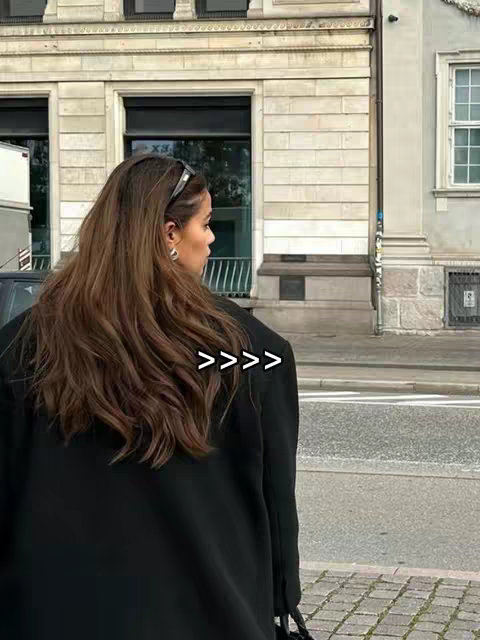A woman with long brown hair is shown from the back, facing towards the left, in front of a building. She wears a black blazer and a silver hairband, with her face partially visible. The background displays a light-colored building and a partially visible street.