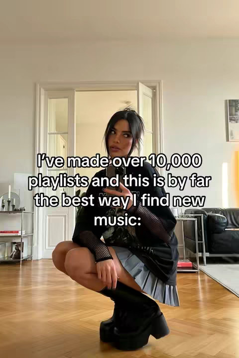 A woman is kneeling in an off-white room, with natural light spilling in. She has black hair and is wearing a black top, fishnet sleeves, a gray pleated skirt, and black platform boots. Text overlayed on the image says, "I've made over 10,000 playlists and this is by far the best way I find new music:"