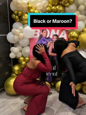Two women, one in a maroon dress and the other in a black dress, are posing in front of a backdrop of gold and white balloons. Both are leaning towards the centre, and a magazine cover with the title 'BONA' on it is visible in front of them. Text overlayed at the top reads 'Black or Maroon?'