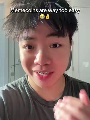 A young Asian man is centered in the frame, looking directly at the camera with a slight smile. Above him, overlaid text reads, "Memecoins are way too easy, " with emojis, while the background appears to be an interior space.