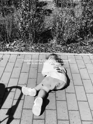 The image is a black and white shot of a child lying on a brick pavement. The child is wearing a t-shirt, shorts, and shoes. They're lying on their stomach with their legs extended and arms at their sides. Behind the child, there are bushes.