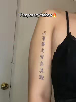 A close-up shot showing a person's upper arm, where a temporary tattoo is visible. The arm is positioned diagonally, with the skin tone appearing fair. The tattoo appears to be a vertical line of Chinese characters running down the arm, while the other arm includes a portion of a black strap.