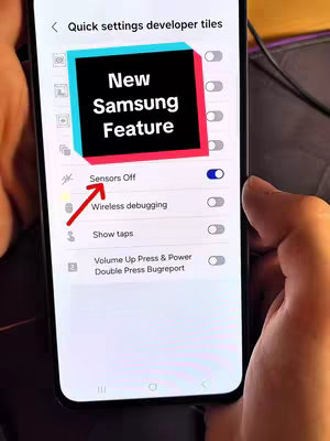 A person's hand holding a Samsung phone, displaying the settings menu. A highlighted section shows 'Sensors Off,' with a red arrow pointing towards it. An overlaid text box reads 'New Samsung Feature'. The color scheme is dominated by the phone's screen against a blurred background.