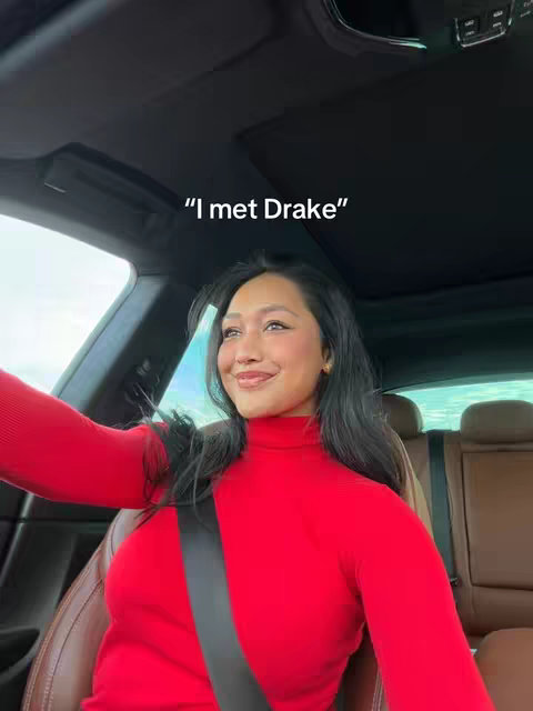 A woman with long black hair and fair skin sits inside a car. She's wearing a red turtleneck. The camera angle is from inside the car, showing her looking at the camera with a slight smile.