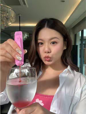 A young woman holding a glass of water with a pink liquid being poured into it from a pink dispenser, with the woman puckering her lips. The background consists of a chandelier and windows.