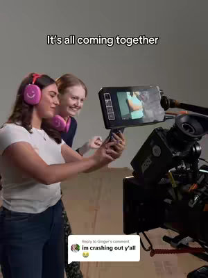 Two young women are standing in front of what appears to be a studio setup. One is wearing headphones with a smiley face on them and seems to be holding a phone up, possibly taking a video. The other woman is smiling looking at what is in front on the video camera with monitor and equipment in the background. The lighting appears to be natural or studio-based.