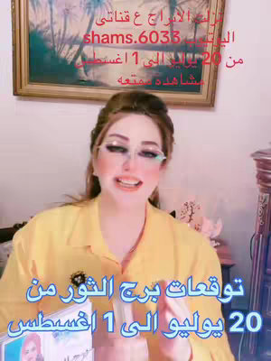 A woman is centered in a selfie-style video, smiling with a pleasant expression. She wears a yellow shirt, has brown hair, and is framed by a background of a framed painting and wooden cabinet. Text overlays in Arabic are placed on the image.