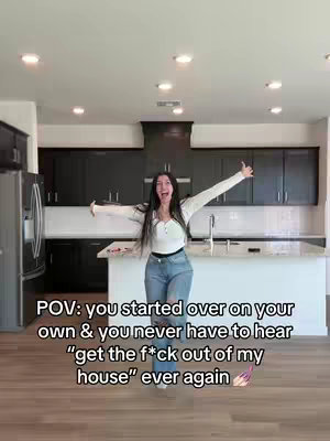A woman stands in a modern kitchen, arms outstretched with a joyful expression. The background includes dark cabinetry, stainless steel appliances, and a light-colored island. Text overlayed reads 'POV: you started over on your own & you never have to hear “get the f*ck out of my house” ever again.'