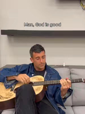 A man is sitting on a gray couch, playing an acoustic guitar. He is wearing a denim jacket and black pants. The text overlay on the image reads, "Man, God is good."