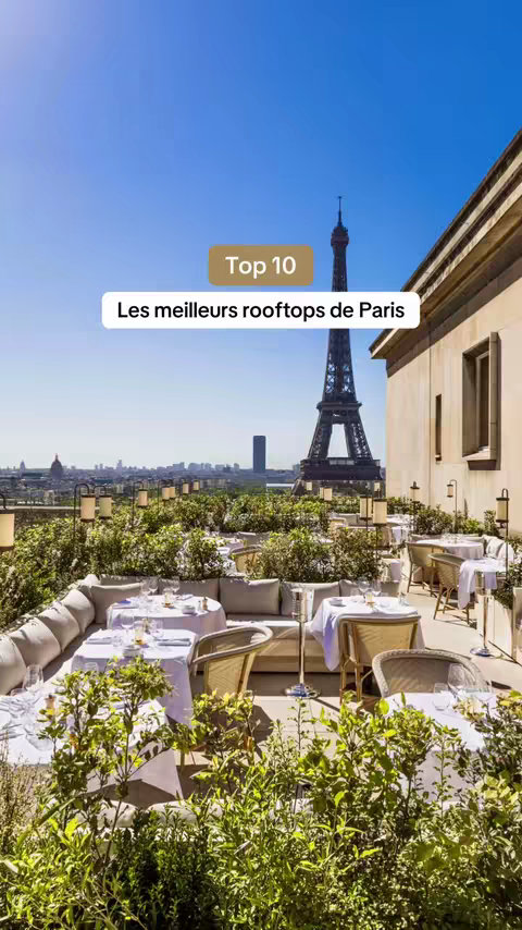 A daytime shot of a rooftop restaurant in Paris, showcasing an exquisite view of the Eiffel Tower. Tables are set with white tablecloths and fine dining settings. Lush greenery, perhaps from plants, provides a natural border around the seating area. The Eiffel Tower dominates the midground, perfectly centered, set under a crystal clear blue sky. The rooftop's structure includes tan-colored walls, windows, and architectural details. The overall layout is well-composed, suggesting luxury and high-end dining.