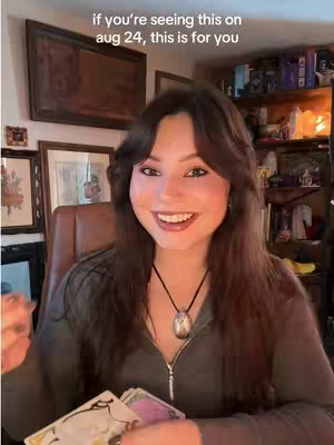 A woman with long brown hair smiles at the camera while holding tarot cards. She wears a zipped-up green jacket and a necklace. Behind her are pictures on the wall and a shelf with items.