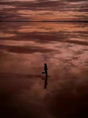 A silhouette of a person is depicted running towards the right in the center of the frame, with a body of water reflecting the cloudy sky above. The ground appears to merge with the sky, creating a mirrored effect. The color palette is dominated by muted oranges, reds, and browns, giving a warm, yet somber, feel. The composition is centered with the horizon line, creating a symmetrical and contemplative mood.