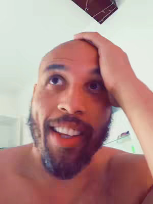 A man with a shaved head and a beard looks up and to the side, touching his head with his hand as if in thought, against a white background. He appears to be smiling or smirking. The angle is from a low perspective.