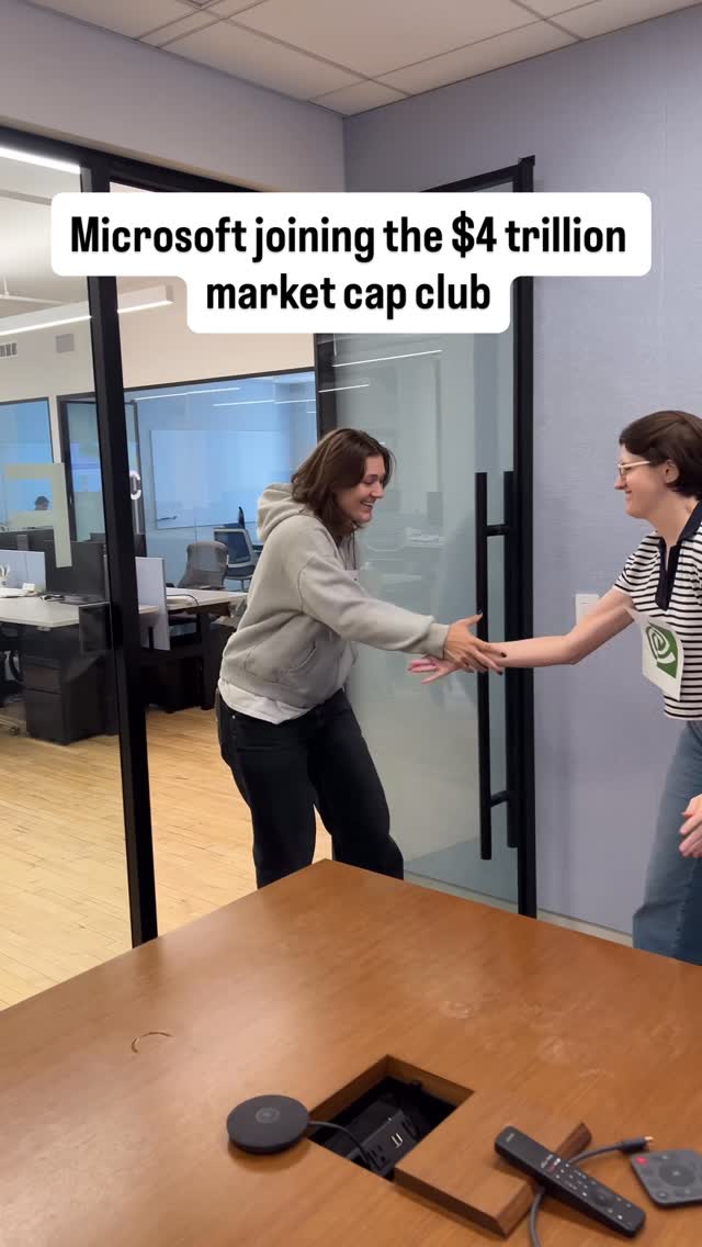 Two women shake hands inside an office setting. The woman on the left wears a gray hoodie and black pants; the woman on the right wears a striped shirt and jeans. A sign that says 'Nvidia' is attached to the shirt of the person on the right. They are standing near a large conference table.