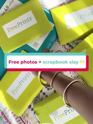 The image shows a top-down view of several wrapped packages of photos arranged on a colorful surface, next to a person's adorned arm, holding the packages. Text overlays read, "Free photos = scrapbook slay".