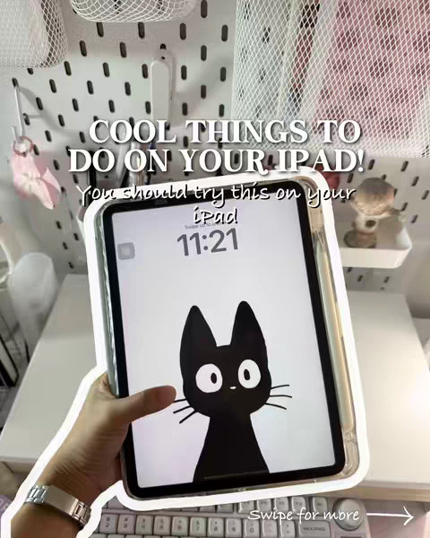 A person is holding an iPad displaying a black cat graphic against a white background. The iPad is in a clear protective case. The background is a white pegboard with accessories, and a keyboard and other items are visible below the iPad. The image is lit with soft ambient light.