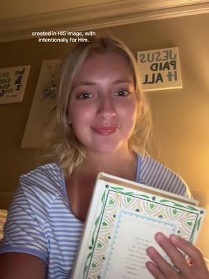 A young woman with blonde hair is holding a book. She is smiling and looking at the camera. The background has some writing on the wall. There is a light source from the top right.