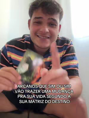 A selfie-style video cover features a young man with short, dark hair, light skin, and a slight smile. He's wearing a striped shirt with black, yellow, and blue stripes. In his left hand, he's holding a tarot card, partially obscuring it. His right index finger is raised, pointing towards the camera. The background is a plain white wall. Text overlay is at the bottom.
