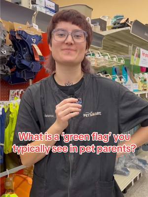 A person wearing glasses and a black PetSmart employee shirt stands in a store aisle. The person is holding a small object near their mouth and is smiling; the store shelves are filled with pet supplies, and an overlay indicates a question about pet parents.