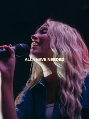 A young woman with long, wavy blonde hair is singing into a microphone. The image is shot from a low angle, looking up at her face, lit with warm tones of pink and red from what appears to be stage lighting, she has a joyful expression. Overlayed in clear white text at the center is the phrase "ALL I HAVE NEEDED". The background is dark, with blurred shapes suggesting an audience or the stage elements.